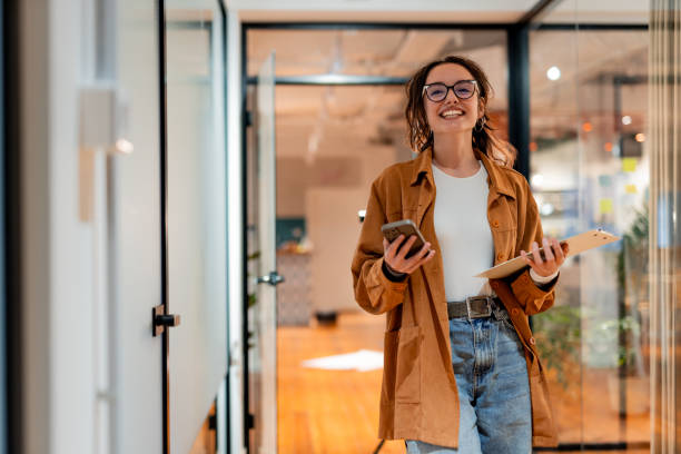istock Young businesswoman walking in office corridor holding smartphone and documents 2196235645
