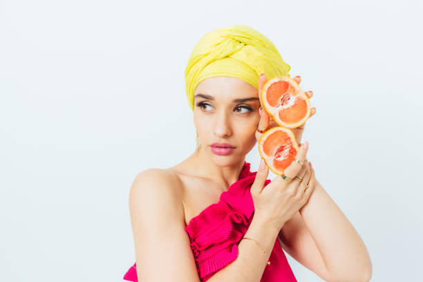 A woman wearing a yellow scarf is holding two oranges. She is wearing a pink dress.