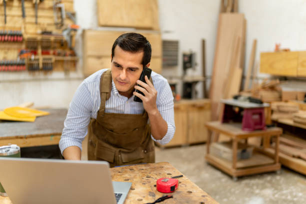 istock Mid adult carpenter man talking on mobile phone and using laptop at carpentry 2196238540