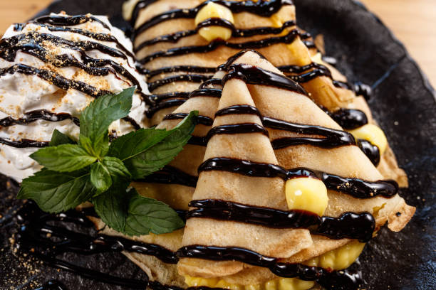 A delectable dessert arrangement featuring crepes filled with custard, alongside whipped cream elegantly topped with chocolate syrup, garnished with fresh mint leaves, and served on a textured black plate