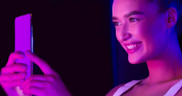 istock Young Woman Smiling While Holding a Smartphone Under Colorful Lights 2196247661