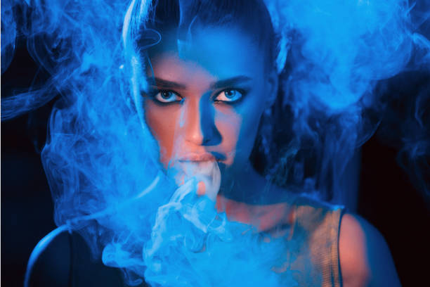 A woman with striking features exhales a cloud of smoke surrounded by vibrant blue lighting, creating a mysterious atmosphere.