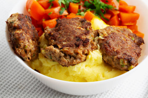 A white bowl with meatballs, mashed potatoes and cooked carrots decorated with parsley on table cloth, close-up