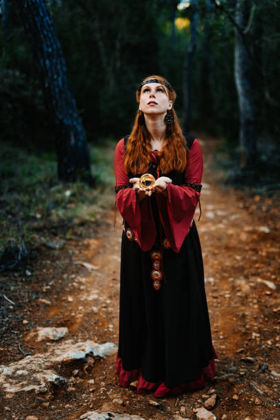 istock Red-haired and beautiful medieval fantasy witch standing in a mysterious forest summoning magic with a golden orb looking up 2196258097