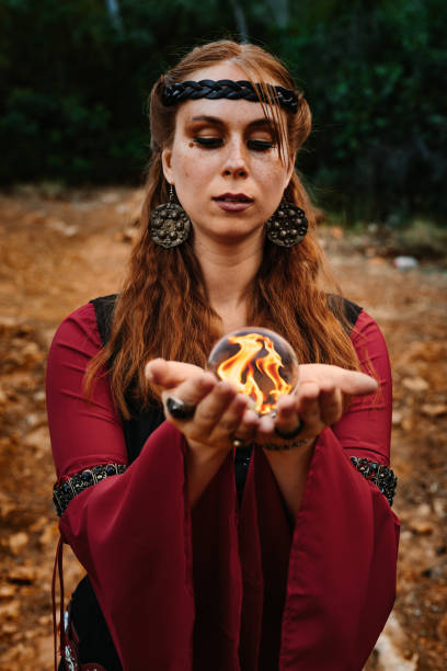 istock Young red-haired and beautiful medieval fantasy witch standing in a mysterious forest summoning magic with a golden orb with fire inside 2196258155