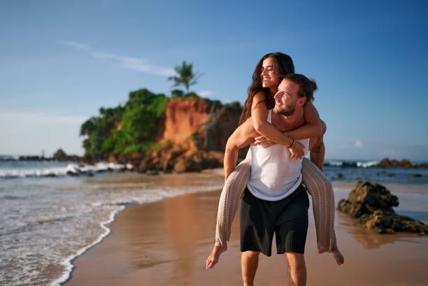 Man gives piggyback ride to smiling woman at sunset. Romantic couple enjoys honeymoon on tropical beach. Newlyweds walk by sea, love moments on exotic island. Happy bride, groom in casual wear. pareja romántica disfruta de luna de miel en una playa tropical. el hombre da un paseo a cuestas a una mujer sonriente al atardecer. los recién casados caminan por el mar, aman momentos en una isla exótica. feliz novia, novio en ropa casual.