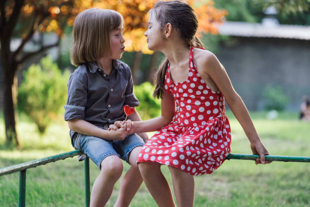 A girl in a polka dot dress playfully holds hands with a boy while enjoying a heart-shaped lollipop as they balance on a green railing in a sunny park setting.