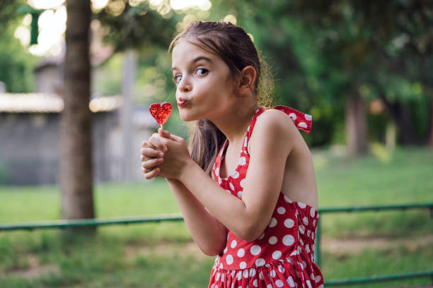 A young girl in a polka dot dress poses affectionately with a heart-shaped lollipop held close to her lips, set against the green backdrop of a park.