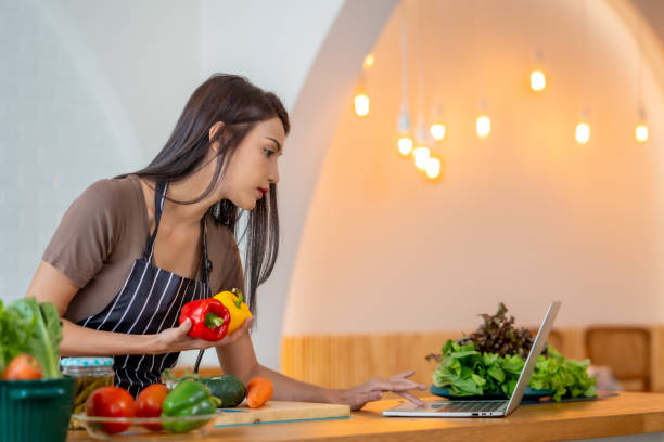 istock woman in apron holds colorful bell peppers while using laptop in modern kitchen. Fresh vegetables and greens are on counter, creating vibrant cooking atmosphere 2196267383