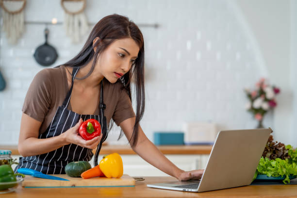 istock woman in apron prepares vegetables in modern kitchen while using laptop. She holds red bell pepper and focuses on screen, showcasing healthy lifestyle 2196267388
