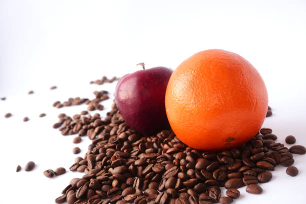 A fresh orange and a red apple rest together on a pile of roasted coffee beans against a white background, combining vibrant colors and textures that contrast the rich brown coffee.