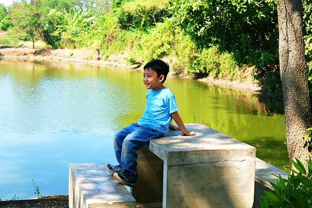 Cheerful young boy sitting on a concrete bench by a tranquil pond, surrounded by lush greenery