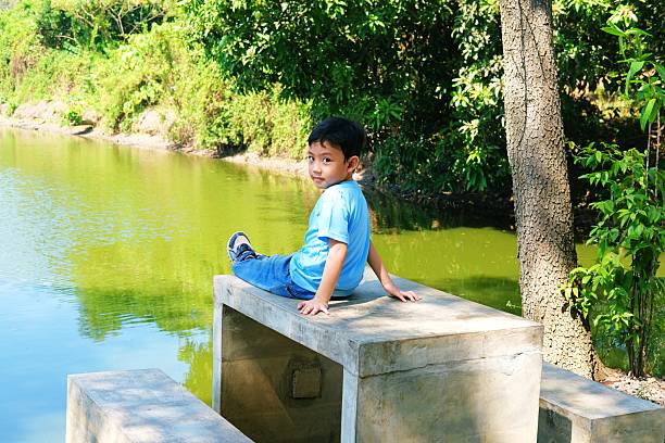 Cheerful young boy sitting on a concrete bench by a tranquil pond, surrounded by lush greenery
