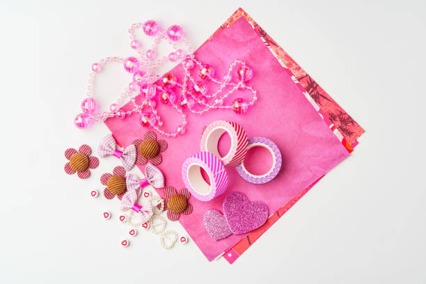 Brightly colored craft supplies lay on a white surface, featuring pink paper, decorative washi tape, beads, flower embellishments, and heart shapes, perfect for various creative projects.