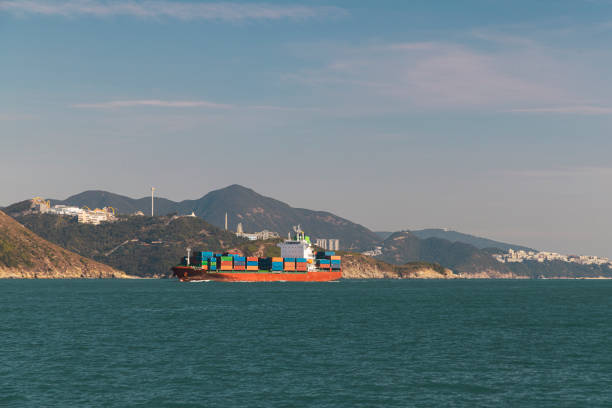 A shipping container vessel moves steadily through Victoria Harbour, surrounded by the scenic landscape of Hong Kong. The mountains rise in the background, showcasing a mix of nature and urban life.