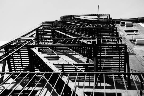 A black and white photograph of a fire escape on a building, showcasing the intricate metal structure and shadows against a clear sky.