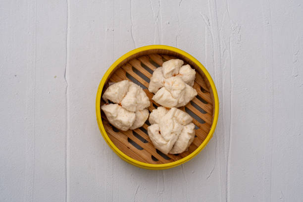 A minimalist top-down view of three freshly steamed char Siu bao (BBQ pork buns) served in a traditional bamboo steamer with a yellow rim. The soft, fluffy texture of the buns contrasts with the rich filling inside. Set against a clean, textured white background, this image captures the simplicity and elegance of classic Cantonese dim sum