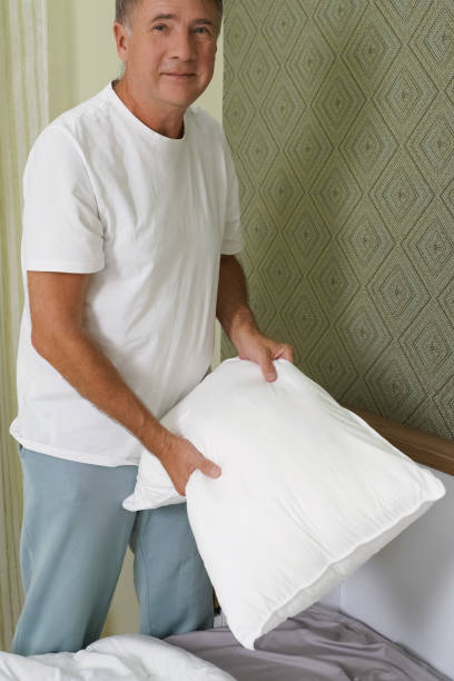 istock A senior man is standing beside his bed, holding a pillow in one hand while looking content. 2196325876