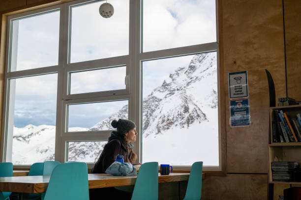Backcountry skier relaxes in ski lodge with hot beverage and looks out to snowcapped peaks, B.C. Coast Ranges