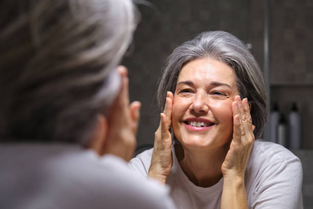 istock Smiling senior woman applying moisturizer cream on face looking at mirror in bathroom 2196339855