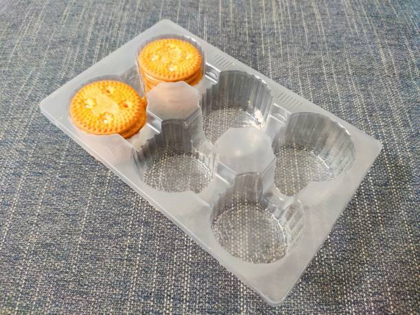 a plastic tray designed to hold cookies or biscuits. The tray has six compartments, but only two compartment is occupied by a stack of round, golden-brown cookies with a patterned surface and small white spots. The tray is placed on a textured, blue fabric surface. This image is relevant for showcasing food packaging, specifically for cookies or biscuits, and can be used in contexts related to food storage, packaging design, or product presentation