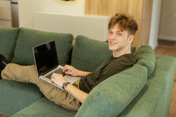 istock Young man working intently on laptop while relaxing on a green sofa in a cozy living room with natural light streaming in 2196358817