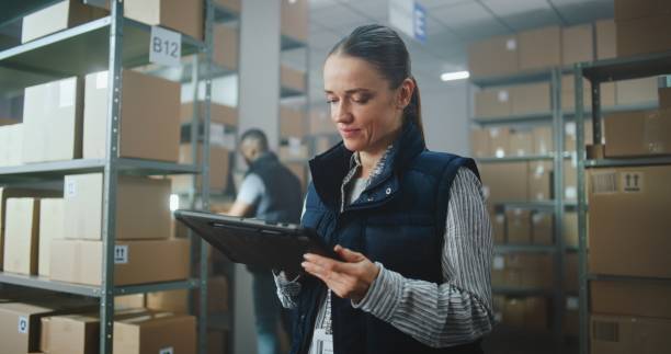 istock Female Inventory Manager in Warehouse Facility: Uses Tablet Computer, Looks at Camera 2196364317