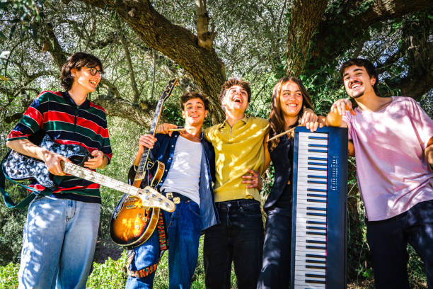 Group of friends from generation z having fun with the musical instruments of their music band, laughing in the shade of a large olive tree