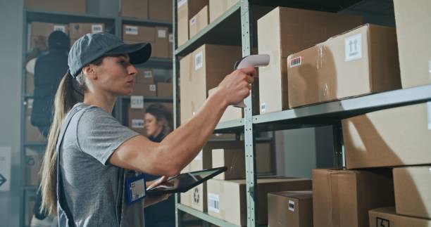 istock Female Warehouse Worker Scans Cardboard Box, Using Tablet Computer, Takes Parcel for Shipping 2196371688