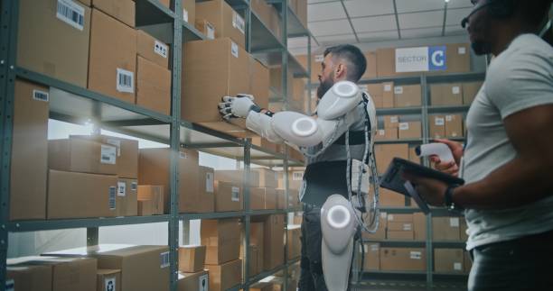 istock Worker Wearing Robotic Exoskeleton Puts Cardboard Box on Shelf. Logistics Specialist Scans Parcel 2196372455
