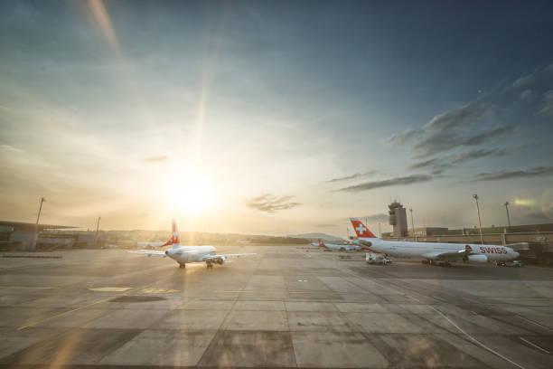 Zurich, Switzerland - September 30, 2023: A breathtaking view of Zurich Airport during sunset, with SWISS aircraft on the tarmac. One Airbus is taxiing towards the runway while another remains parked at the gate, preparing for its next journey. The golden hues of the setting sun cast a warm glow over the airport, creating a serene yet dynamic atmosphere. The control tower stands tall in the background, overseeing the bustling air traffic. This editorial image captures the essence of Swiss aviation, efficiency, and international connectivity.