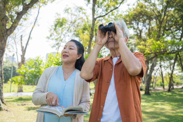 A senior couple enjoying a leisure activity outdoors in a public park. The senior man using binoculars to observe birds in the distance while the senior woman holding an open book standing side by side with him engaging in a discussion.