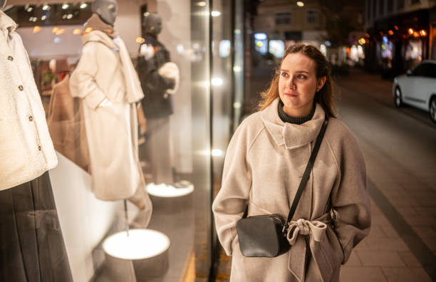 Young woman with hands in her pockets of coat walking on the street and looking at windows store at night