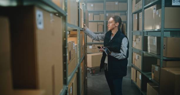 istock Female Warehouse Associate Scans Cardboard Boxes, Uses Tablet Computer, Takes Parcel from Shelf 2196396523