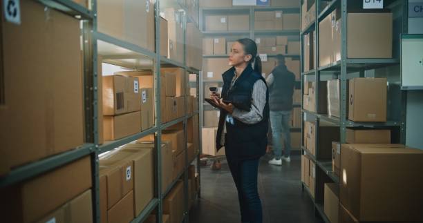 istock Female Warehouse Associate Scans Cardboard Boxes, Uses Tablet Computer, Takes Parcel for Shipping 2196400695