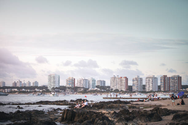 A tranquil sunset in Punta del Este, where the golden light bathes the city's modern skyline.\nIn the foreground, people unwind along the rocky shoreline, embracing the peaceful coastal atmosphere. Meanwhile, the bay comes to life with yachts and boats gently drifting on the water.