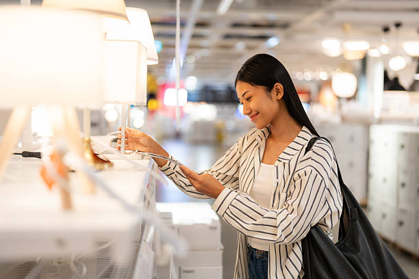 Young Asian woman browsing and testing light fixtures in a well-lit home decor store, representing home improvement, interior design choices, and a modern consumer shopping experience.