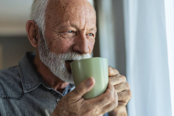 istock A joyful elderly man with a beard stands by a window, holding a green cup in his hand, exuding warmth and happiness in a beautifully lit, modern living space. 2196403459