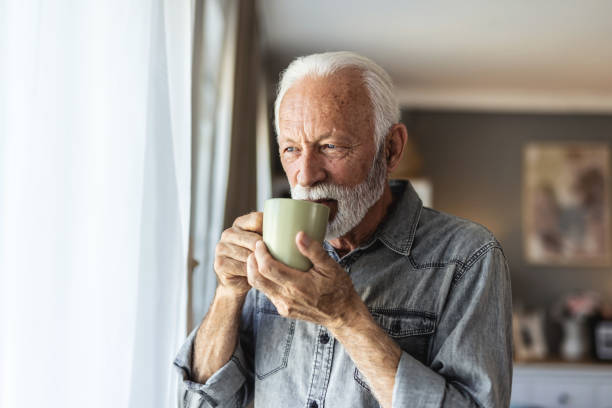 istock A joyful elderly man with a beard stands by a window, holding a green cup in his hand, exuding warmth and happiness in a beautifully lit, modern living space. 2196403518