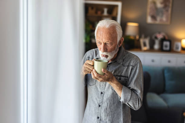 istock A joyful elderly man with a beard stands by a window, holding a green cup in his hand, exuding warmth and happiness in a beautifully lit, modern living space. 2196403553