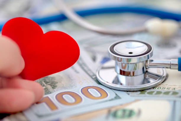 Close-up of a hand holding a red heart. Stethoscope on a heap US $100 dollars bills in the background. Shallow depth of field.