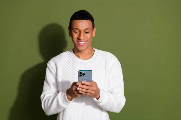 istock Young man in white sweater smiling while using smartphone against khaki background 2196410481