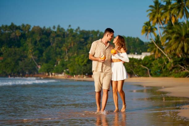istock Romantic couple on beach holiday stroll hand in hand, sipping fresh coconuts. Laughing, chatting, they enjoy golden sunset. Perfect scene for honeymoon, dating app promos, tropical retreats. 2196413825