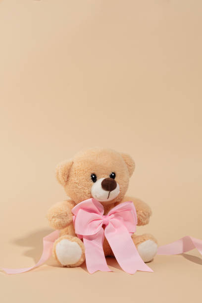 A plush teddy bear with a cheerful expression, adorned with a pink ribbon, set against a warm beige backdrop ideal for Christmas illustrations