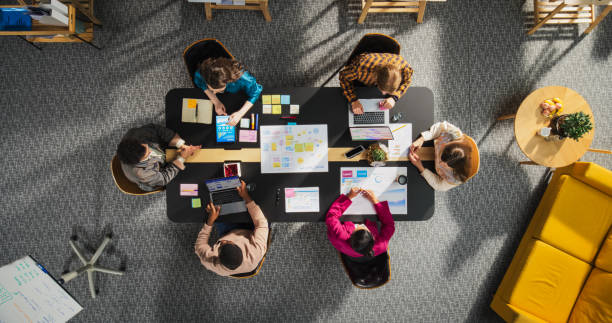 Top Down View of Young, Diverse Entrepreneurs in a Colorful Office, Solving Problems and Designing Software, Using laptop, Tablets, and Creative New Project at Conference Table. Productive Teamwork