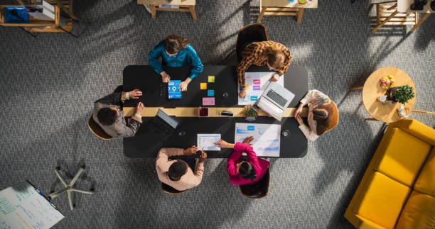 istock Startup Meeting Room: Team of Entrepreneurs Sitting Around Conference Table, Having Discussions, Solving Business Problems, Using Tablet, Laptop, Share Documents with Statistics, Charts. Top Down View 2196418515