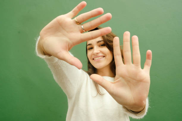 A young woman in a white sweater smiles joyfully while playfully reaching toward the camera. She stands in front of a vibrant green wall, creating a striking contrast. Her relaxed pose and expressive hands add a fun, dynamic energy to the image.