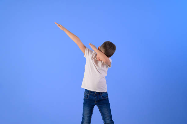istock Little carefree boy with arms raised dancing and dabbing while standing on isolated blue background 2196426627