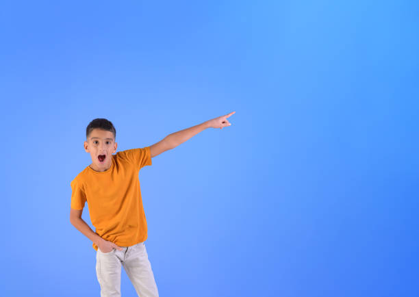 istock Portrait of surprised boy with mouth open pointing at blank space while standing on blue background 2196426882