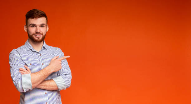 istock Young Man Gestures Towards Empty Space on Orange Background During Informal Photoshoot 2196431540